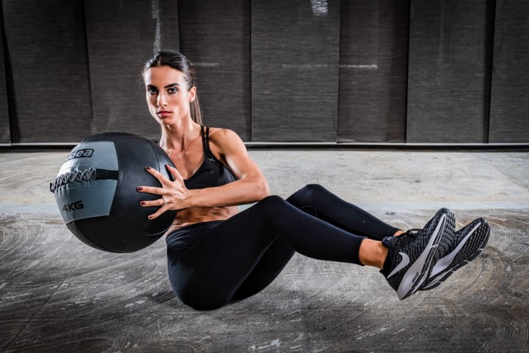 A woman holding a wall ball fitness equipment laying down on the floor of a gym.