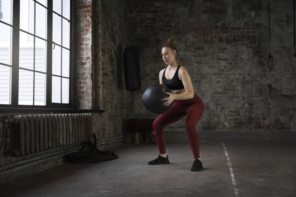 A woman holding a wall ball fitness equipment trying to do squats. 